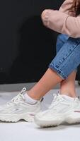 Cropped image of a girl sitting on a black step. Model in jeans, sweatshirt and white sneakers in studio. Vertical video