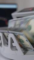 Switching on cash counting machine to count a wad of one hundred dollar bills. Close up. Laptop at backdrop in blur. Vertical video