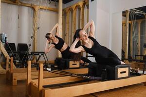 Two women in black sportswear are doing pilates on a reformer machine in the gym photo
