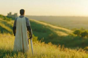Knight standing on a grassy hill at dusk with a sword across his shoulders and a serene sunset backdrop photo