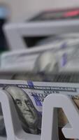 Notes with Benjamin Franklin portraits fall on the holder of cash counting machine. Close up. Blurred background. Vertical video