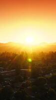 Beautiful cozy green city in the bright warm rays of setting sun. Sun hiding behind the mountains. Orange sky at backdrop. Aerial view. Vertical video