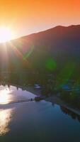 Last rays of setting sun peeping from behind the high mountains. Beautiful lake with urban infrastructure at the shore. Orange sky at backdrop. Vertical video