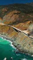 Idyllic scenery of turquoise ocean and crushing waves at the rocks. Highway passing through the mountains. Sunny day backdrop. Vertical video