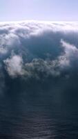 Blue ocean water covering with thick fog. Bright sun shining on the top of hazy cloudscape. Aerial view. Vertical video