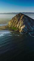 Flying closer to a pyramid-shaped mountain at the coast of Pacific. Little white waves coming slowly to the shore. White fog covering the horizon. Vertical video
