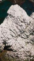 White rock peeping out from blue water of ocean on sunny day. Waves hitting the mount. Seaweeds floating on the surface of water. Top view. Vertical video