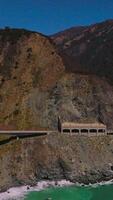 Road connecting the mountains at the coastline of California. Beautiful sight of the rugged rocks at the backdrop of blue clear sky. Vertical video