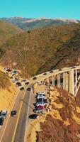 Cars moving slowly to and from the bridge in the mountains. California rocks at the coast of Pacific on sunny daytime. Aerial view. Vertical video