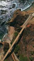 View of the roads in the mountains connected by the bridge. Rocky coastline of California with beautiful blue ocean from aerial perspective. Vertical video