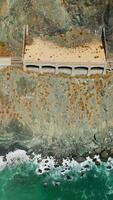Road through the mountainous shore covered with shade in one part. Foamy waves crushing by the rocky coastline. Aerial perspective. Vertical video