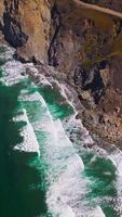 Aquamarine ocean waters with white foamy waves splashing by the rocks. Some houses and motorway are on the top of the mountain. Aerial view. Vertical video