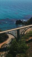 Sightly arched bridge for transport between the mountains on the coast of Pacific. Beautiful blue water sparkling in the sun at backdrop. Vertical video