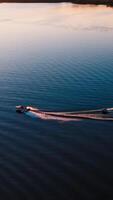 Motor boat on water at sunset. Panoramic view of the evening river with two boats. Motor boat pulling small boat with people. View from above. Vertical video
