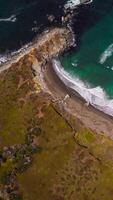 Flying over the rocky shore and moving to the azure waters of ocean. Little white waves splashing by the mountainous coastline. Top view. Vertical video