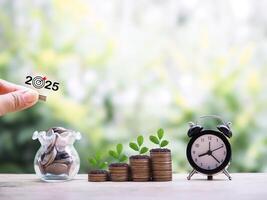 Hand putting coin into glass bottle, Black alarm clock and Plants growing up on stack of coins. The concept of saving money, Financial, Investment and Business growing up in year 2025. photo