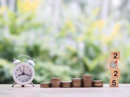 White alarm clock, Wooden blocks with the number 2025 and stack of coins. The concept of saving money for manage time to success business in year 2025 photo