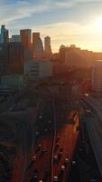 Multiple cars move by the roads and stand on the parking lots. Beautiful downtown skyscrapers of Los Angeles in the rays of setting sun. Vertical video
