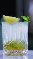 Misted glass full of beverage with two short black straws in it. Rotating glass with lemon and mint leaves on top. Close up. Blurred backdrop. Vertical video