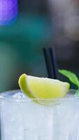 Upper part of glass filled with light beverage and two black straws in. Ice cubes, lemon slice and leaves of mint on top. Close up. Blurred backdrop. Vertical video