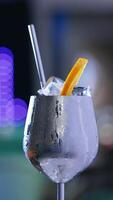 Rotating metal wineglass with misted surface. Cocktail with ice and orange slice on top and metal straw inside. Close up. Blurred backdrop. Vertical video