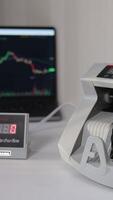 Man puts a pack of one hundred dollar banknotes into a cash counting machine. US bills counted in equipment. Laptop with chart at backdrop in blur. Vertical video