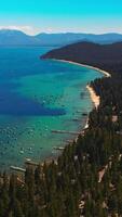 Turquoise waters of amazingly beautiful Lake Tahoe with wooded shore. The Sierra Nevada mountains at backdrop. Aerial view. Vertical video
