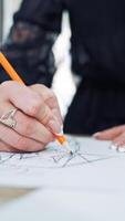Hands of a fashion designer with a beautiful manicure draw a sketch with a pencil. Professional dressmaker creates a stylish design of a new dress on a sheet of paper. Close-up Vertical video