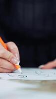 Hands of a professional tailor is drawing a sketch with a sharp pencil on a table. Dressmaker is making a creative design of a dress on a sheet of paper in her workplace. Vertical video