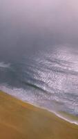 Sandy beach with waves moving to it. Drone footage over the coastline and ocean waters. Thick fog covering the horizon. Vertical video