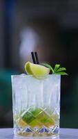 Beverage in old-fashioned glass decorated with lemon and mint leaves. Cocktail with two straws on the rotating table. Blurred backdrop. Close up. Vertical video