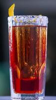 Misted long glass with red drink. Rotating table with cocktail decorated with orange peel. Close up. Blurred backdrop. Vertical video