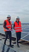 Installation of solar panels. Two workers in protective uniform and hard hats walking along the solar panels on the new photovoltaic station. Vertical video