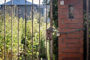 Rusty old lock hangs on a chain at the gate on a red brick post on abandoned territory with greenery photo