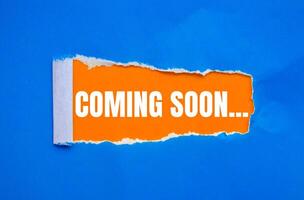 Coming soon message written on ripped torn blue paper with orange background. Conceptual coming soon symbol. Copy space. photo