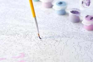 Coloring by numbers. close-up of a paintbrush hovering above a numbered canvas with paint pots in soft pastel colors. photo