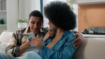 Happy married African American couple man and woman at home couch looking mobile phone shopping website app store internet using smartphone search booking tickets choosing purchase discussing talking photo