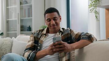 African American guy ethnic happy smiling man relaxing on comfortable couch sofa at home using mobile application scrolling phone app chatting social network online web surfing internet smartphone net photo