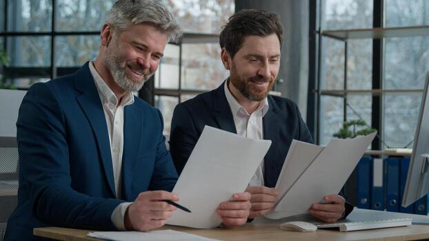 Two Caucasian men businessmen in office working together on financial statistics analyzing charts project discussing data in report doing paperwork using computer looking documents client consultation photo