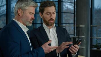 Executive Caucasian men businessmen team business partners colleagues working smartphone on financial statistics analyzing data in report discussing project results discussing business phone office photo