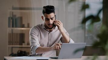 Busy millennial arabian business man web designer employee using laptop talking on cell phone involved in multitasking using gadgets discussing online project distant noting paper documents at office photo