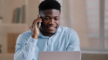 Smiling african man male businessman employee freelancer multitasking worker sit at home office talk on cellphone look at laptop screen provide remote communication business call use computer service photo