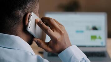 Backside unknown african american man young businessman web designer look at laptop screen surf internet website scroll page interface in social network talk mobile phone wireless call communication photo