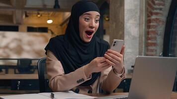 contento emocionado alegre árabe musulmán negocio mujer preguntarse asombro hembra islámico niña hijab usuario estudiante mujer de negocios móvil teléfono bueno Noticias lograr victoria en línea apuesta conmoción ganar teléfono inteligente oficina foto