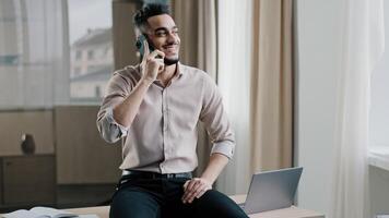 Smiling arab man relaxed male entrepreneur businessman sitting top of table in workspace holding negotiation distant cellphone call with client conversation discussing with colleague by mobile phone photo