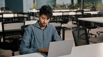 Young man Arabian employee writer thoughtfully typing laptop keyboard copying text in electronic form focused man checking original documents writing new article for website checking figures in report photo