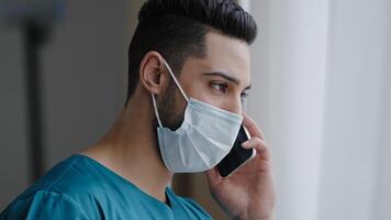 Latin man medical assistant doctor in medical mask consulting patient remote discussing issues by phone hispanic adviser using mobile healthcare app standing near window distant consultation concept photo