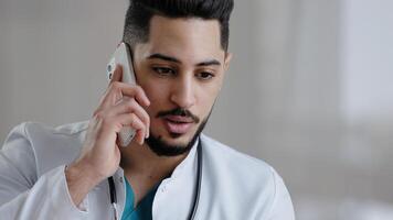 Serious male hispanic guy doctor therapist medical assistant consult patient distant discuss issues by mobile phone arab man adviser surgeon provide remote consultation use cellphone at clinic cabinet photo