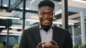 Happy smiling african guy american man businessman boss employee holding modern wireless device texting message on smartphone using social media apps browsing internet playing mobile game at office photo
