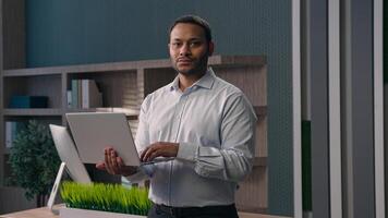 American male office worker standing at workplace in corporate company businessman working with computer man work online typing chatting with clients business task holding laptop smiling at camera photo